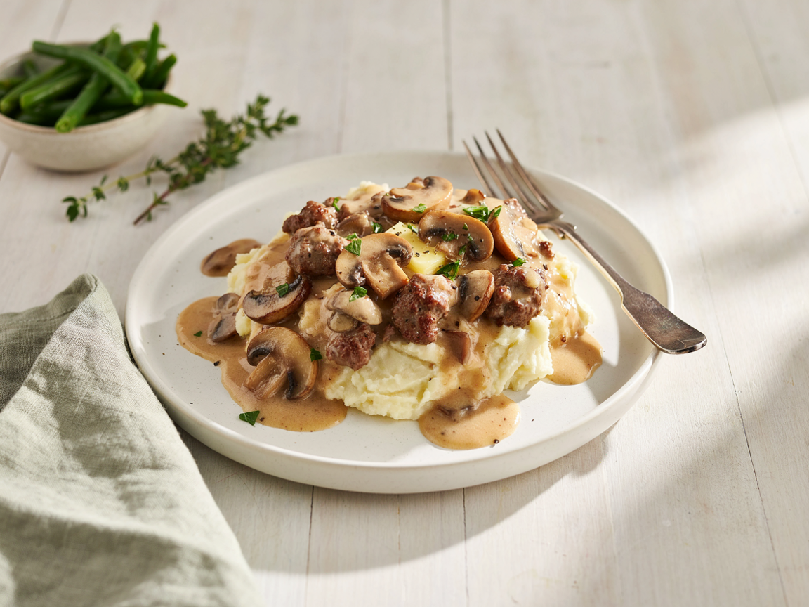 Hamburger stroganoff with mushrooms spooned over creamy mashed potatoes on a white plate, with green beans and thyme on the side