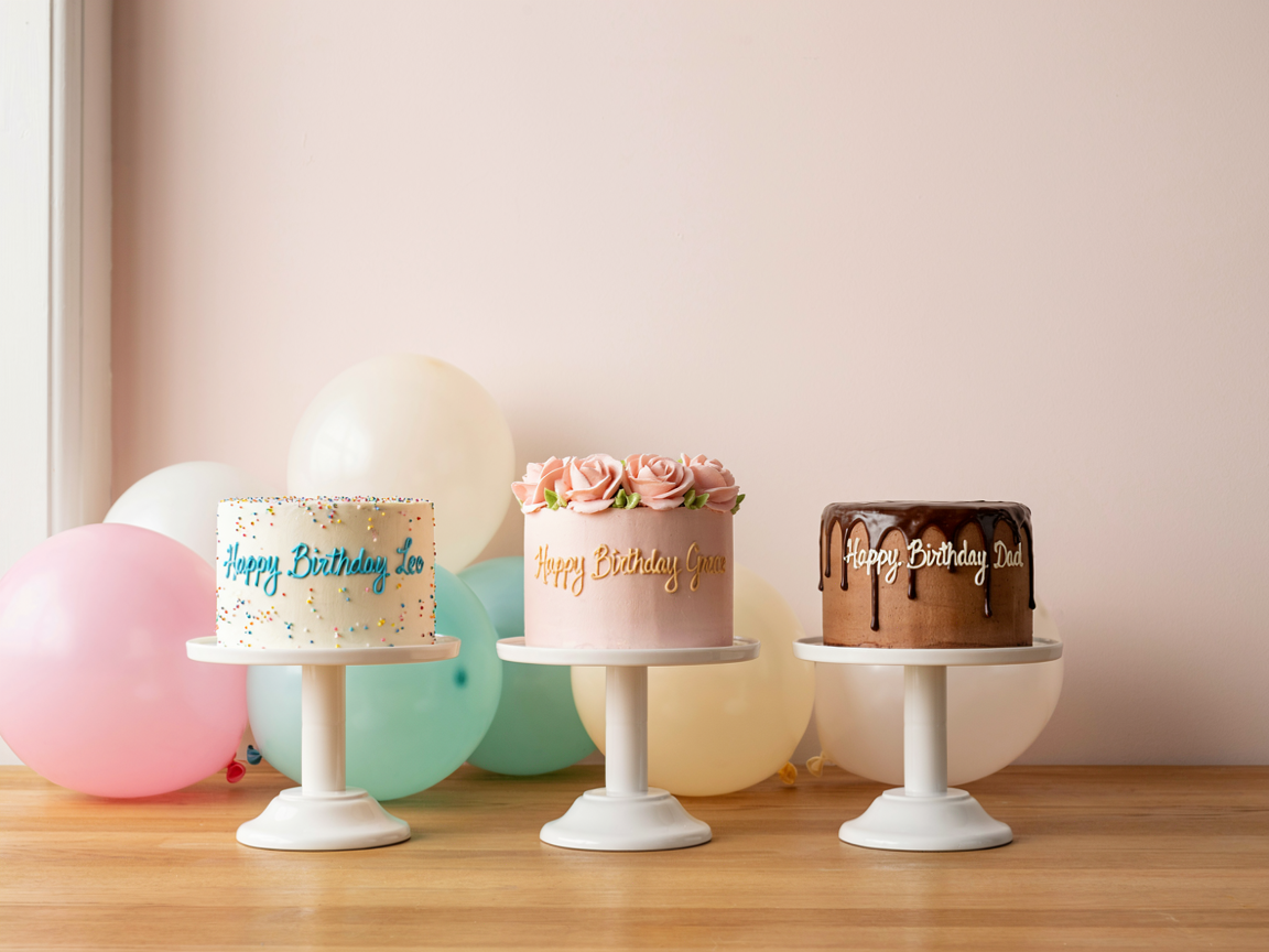 Three small birthday cakes on white stands, each piped with a different name, with pastel balloons behind