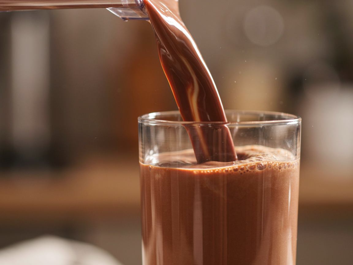 Thick chocolate protein shake being poured into a tall glass with a cream-coloured layer swirling through it