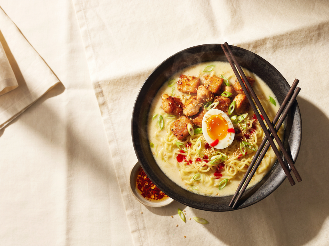 Spicy fiery chicken ramen with creamy garlic sauce in a dark bowl, topped with crispy chicken, a soft-boiled egg, scallions and chili oil