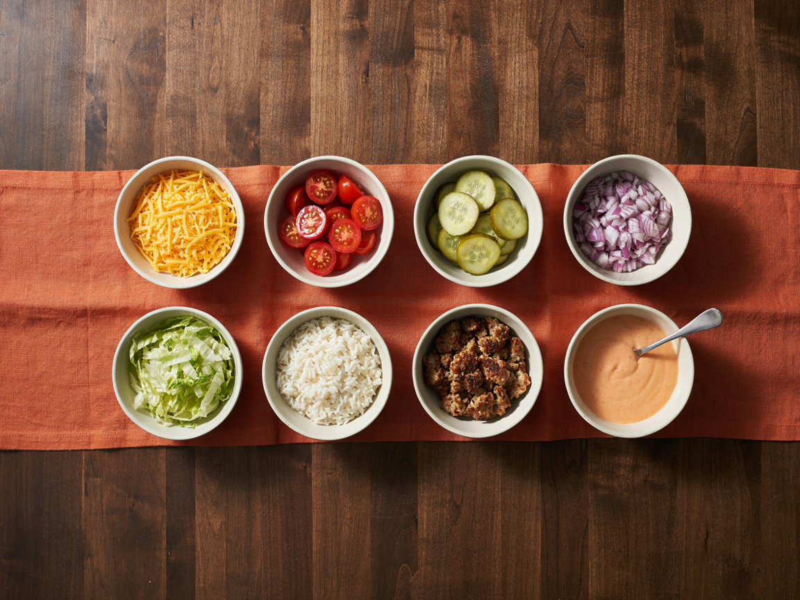 Overhead view of small bowls of toppings including shredded cheddar, tomatoes, pickles, onion and lettuce