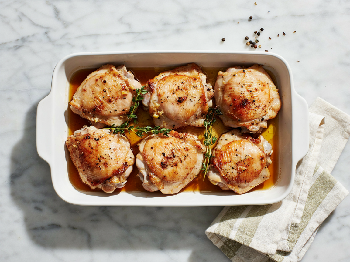 Six seasoned boneless skinless chicken thighs baked golden in a white ceramic dish with thyme and pan juices
