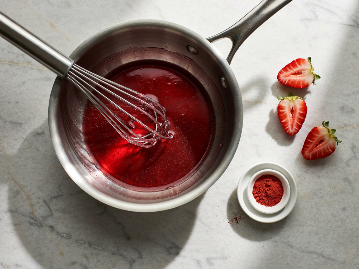 A saucepan of clear glossy red strawberry glaze being whisked, translucent and thickened