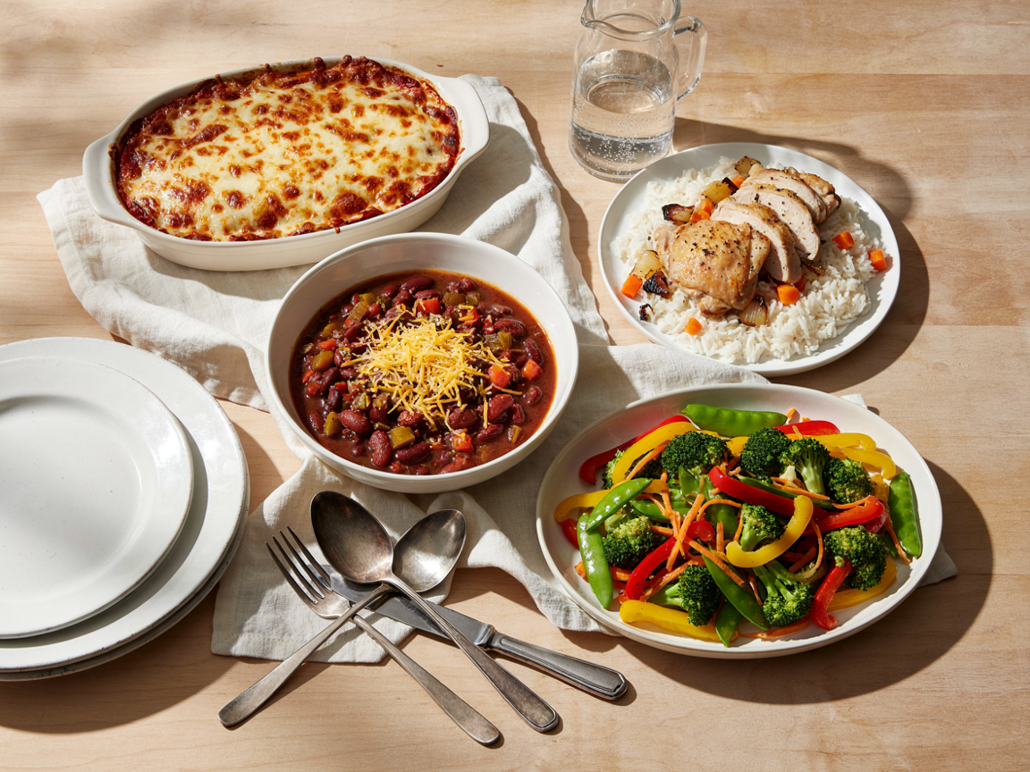 Family dinner table set with four budget meals including baked pasta, one-pan chicken and rice, bean chilli and a vegetable stir fry