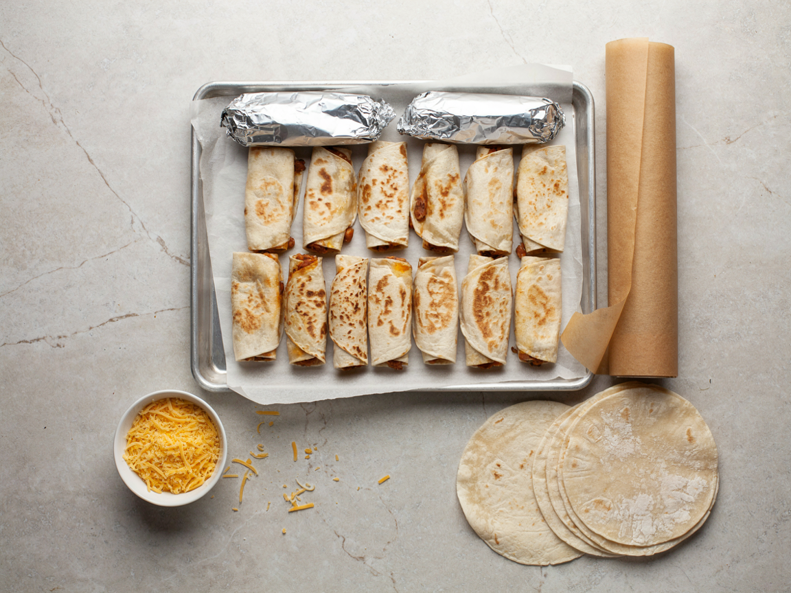 Sheet pan of rolled bean and cheese burritos on parchment with two foil-wrapped burritos ready for the freezer