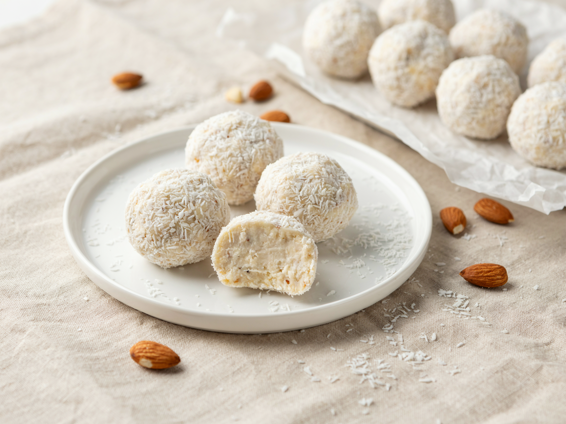 White plate of coconut protein balls rolled in shredded coconut, one bitten to show the creamy centre, more on parchment, almonds and coconut flakes, beige linen