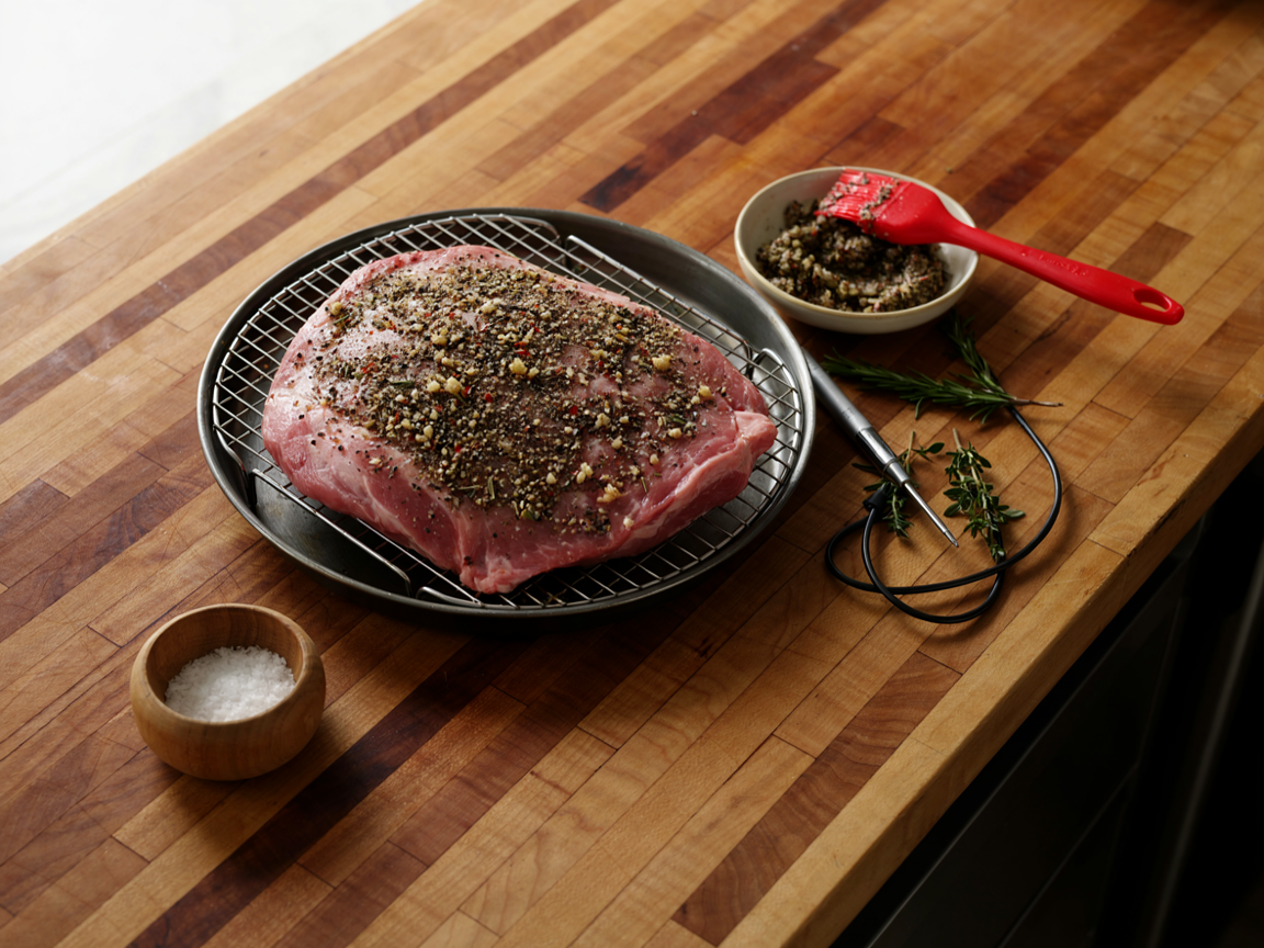 A raw eye of round roast on a wire rack in a roasting pan being coated in a dark herb, garlic and coarse pepper paste, a small bowl of the paste with a brush, sprigs of rosemary and thyme, a bowl of kosher salt and a probe thermometer beside the pan on a butcher block counter