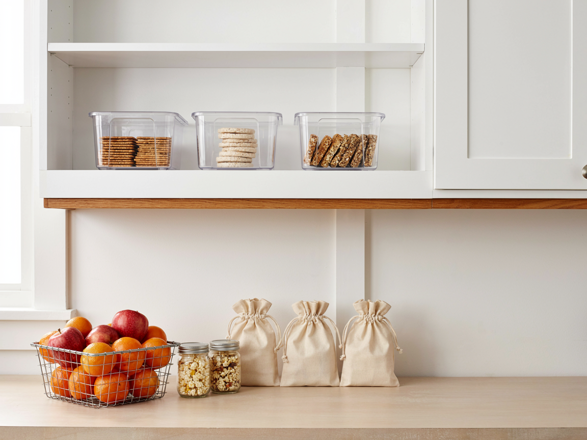 Organized pantry snack station with clear bins of crackers and snacks on a white shelf, a basket of apples and clementines and jars on the counter