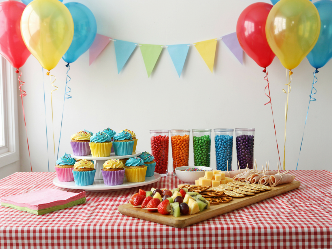 A kids birthday party food table against a white wall: a tiered stand of cupcakes with sky-blue and yellow frosting, a row of clear cups filled with colorful candy, a wooden grazing board of cheese cubes, crackers, rolled turkey bites and fruit skewers in front, red, yellow and teal balloons, paper bunting, red gingham cloth, bright cheerful light