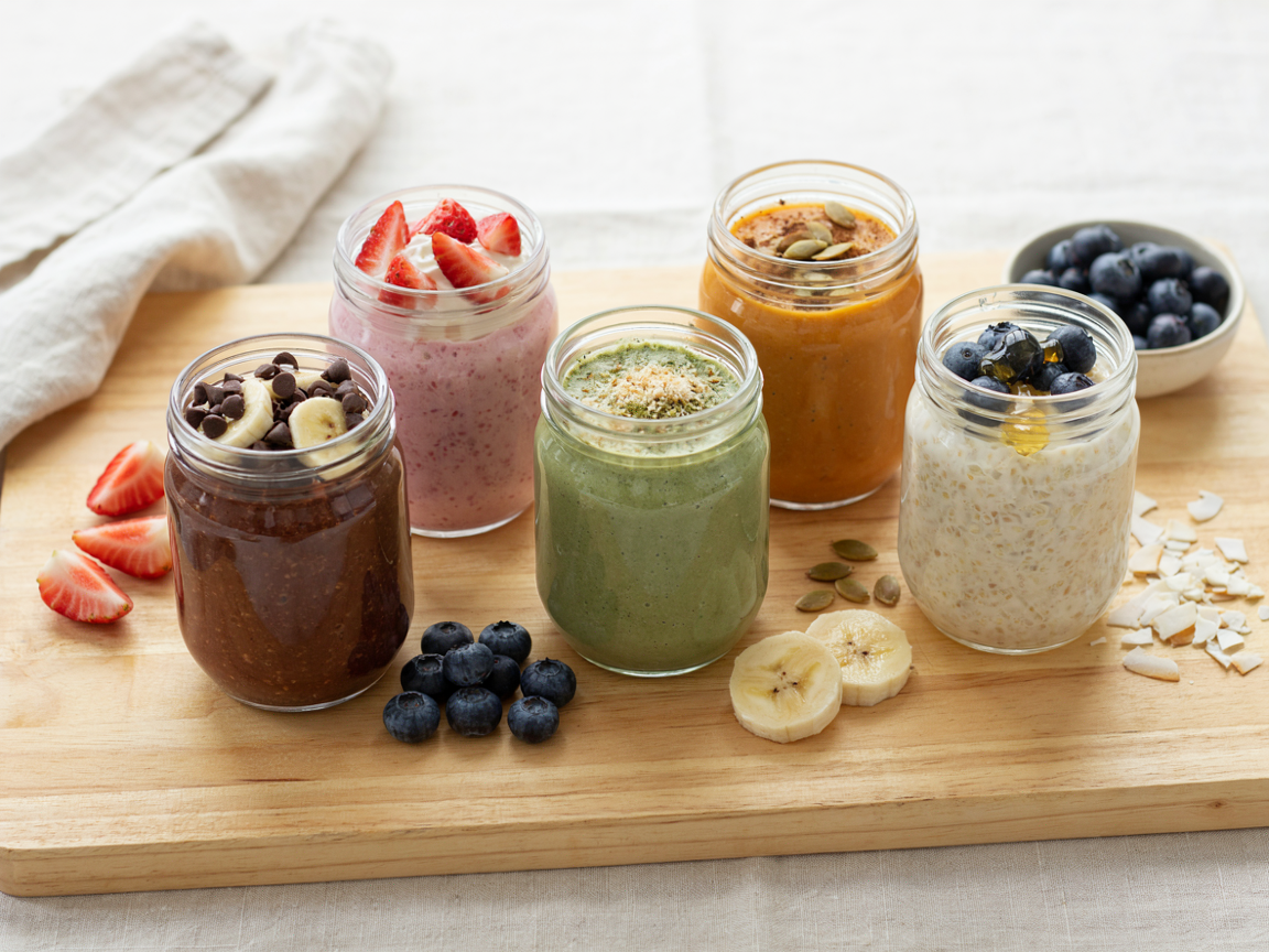 Five jars of blended overnight oats lined up on a light wooden board: a chocolate brownie jar, a pink strawberry cheesecake jar, a pale green matcha jar, an orange pumpkin spice jar and a vanilla jar topped with blueberries, soft daylight