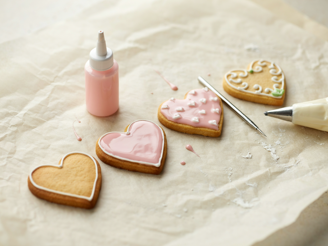 Close-up of sugar cookies being decorated with royal icing: one outlined, one flooded, one with wet-on-wet dots, one with piped details, a piping bag and scribe tool beside them