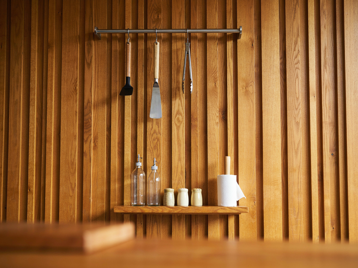 Close-up of a slatted cedar back panel with a stainless rail holding a spatula, a scraper and tongs on hooks, and a small cedar spice shelf with unlabeled bottles