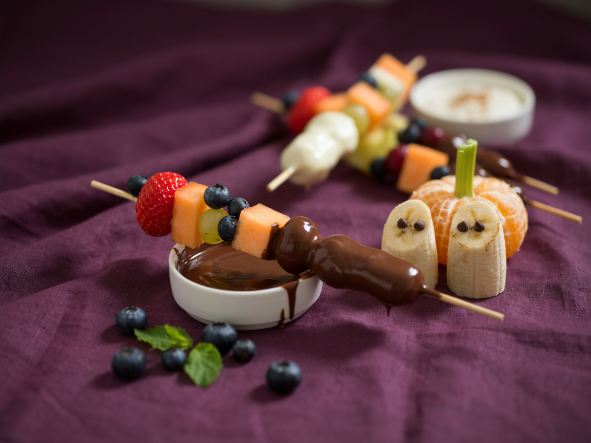 Close-up of Halloween fruit skewers with strawberries, green grapes, cantaloupe and blueberries, one dipped in chocolate, beside banana ghosts with chocolate chip eyes and clementine pumpkins