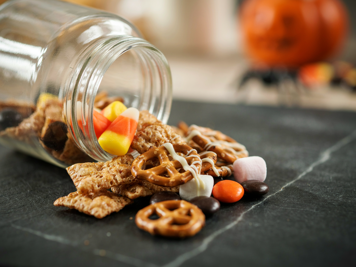 Close-up of Halloween snack mix spilling from a glass jar: caramel cereal clusters, pretzels, marshmallows, candy corn and orange and black candies