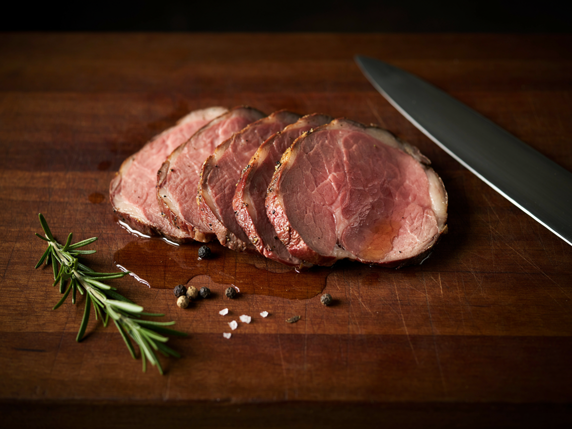 Close-up of thin slices of eye of round roast beef fanned on a board, each slice blush pink from edge to edge with a thin dark peppery crust, juices pooling, a carving knife blade and a sprig of rosemary at the edge