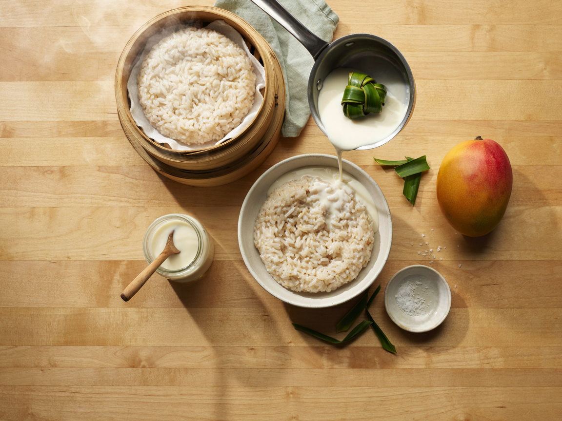 A kitchen counter from above: a bamboo steamer basket lined with cloth holding freshly steamed glutinous rice, a ceramic bowl of hot rice with warm coconut cream being poured over it from a small saucepan, a knotted pandan leaf in the pan, a small glass jar of coconut cream, a whole ripe mango and a small dish of salt, soft natural light