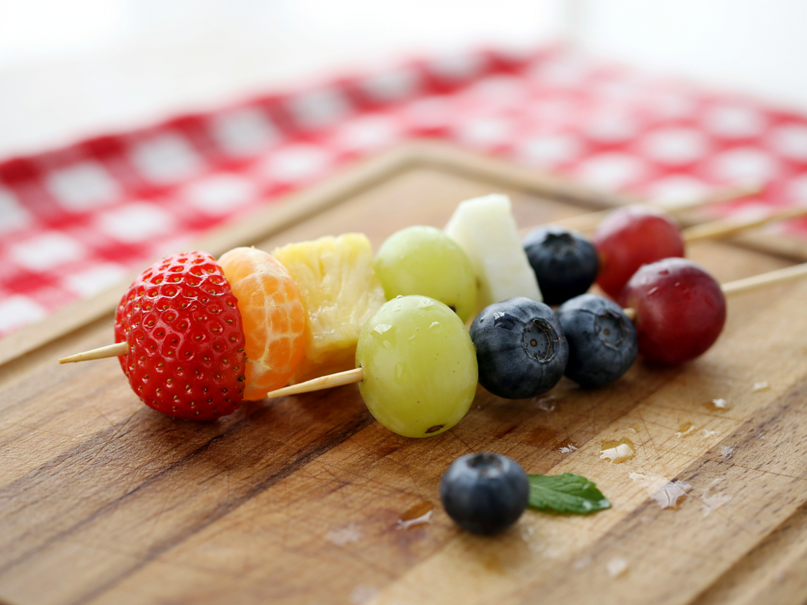 Extreme close-up of rainbow fruit skewers on a wooden board: a red strawberry, an orange mandarin segment, a yellow pineapple chunk, a green grape, three blueberries and a red grape on each, glistening, red and white gingham blurred behind, bright natural light, shallow depth of field