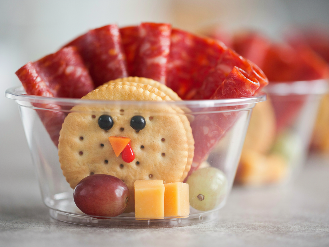 Extreme close-up of a single turkey charcuterie cup: a round golden butter cracker face with two candy eyes, a tiny orange cheddar triangle beak and a red pepper wattle, thin red salami slices fanned behind like feathers, a red grape and cheddar cubes in front, clear cup, soft light, shallow depth of field