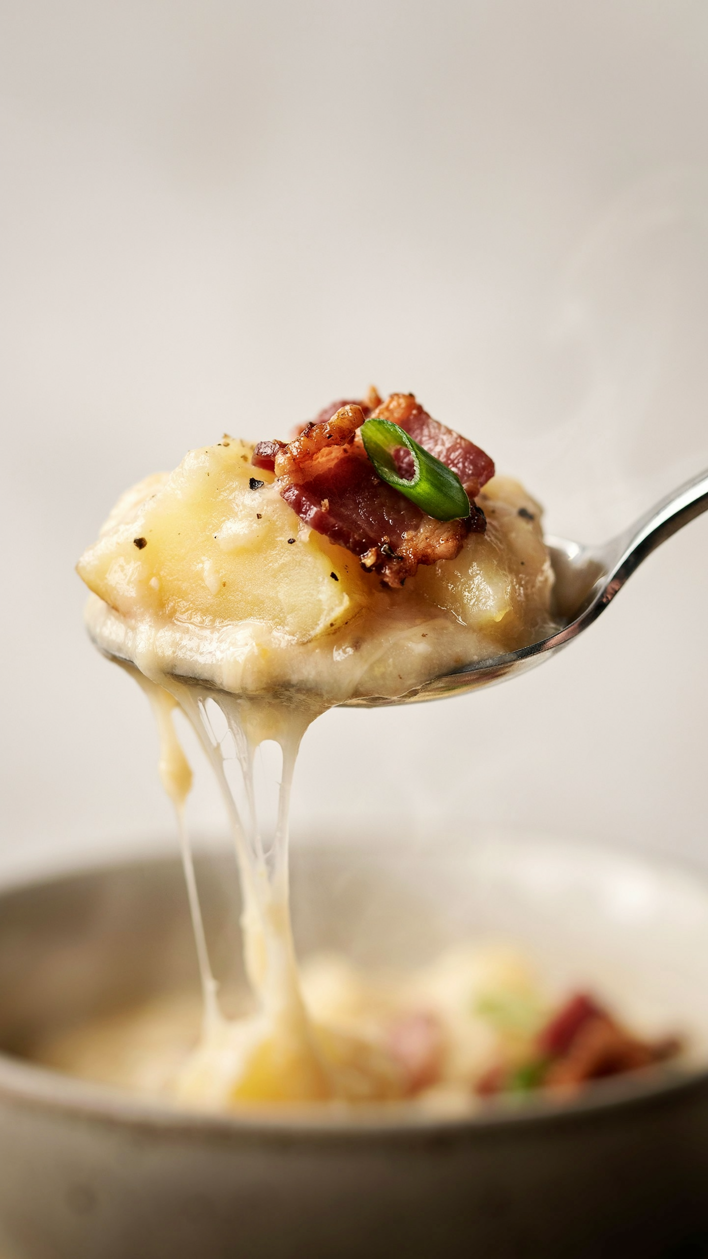 Extreme close-up macro of a spoonful of loaded potato soup lifted from a bowl: thick creamy pale-gold soup coating the spoon, a chunk of tender potato, strands of melted sharp cheddar stretching, a crumble of crispy beef bacon and a sliver of green onion, soft directional light, shallow depth of field
