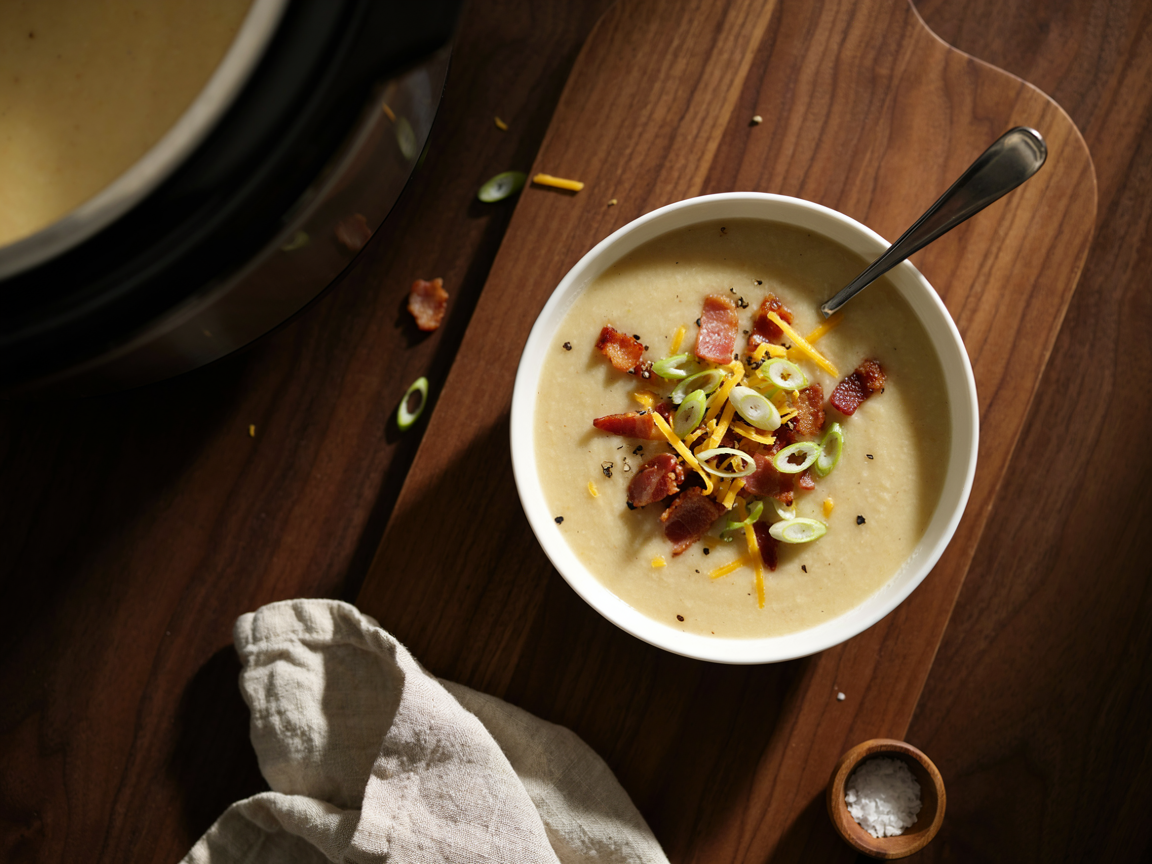 A white ceramic bowl of ultra-creamy potato soup seen from above, a pale velvety soup topped with crispy beef bacon bits, golden dairy-free cheddar shreds, sliced green onions and cracked black pepper, a spoon resting in the bowl, on a wooden board with a linen napkin, a slow cooker softly blurred at the edge, warm autumn light