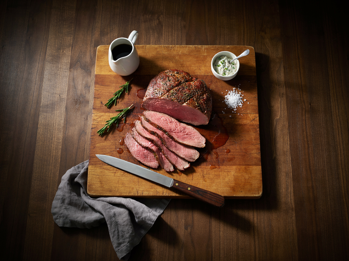 Poor man&rsquo;s prime rib on a wooden carving board: an eye of round roast with a dark mahogany herb and pepper crust, half sliced into thin blush-pink medium-rare slices fanned forward, a small pitcher of au jus, a bowl of horseradish cream, fresh rosemary and coarse salt on a dark wooden table