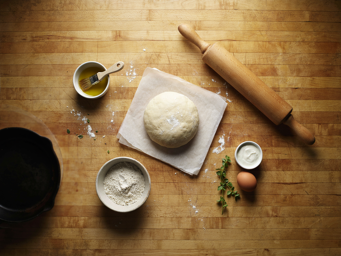 A smooth round ball of pale gluten-free pizza dough resting on oiled parchment on a wooden board, a rolling pin, a small bowl of olive oil with a brush, a bowl of gluten-free flour, a dish of Greek yogurt and a brown egg beside it, a cast iron pan in soft focus behind