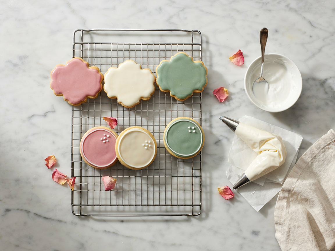 Decorated sugar cookies flooded with pink, cream and sage green royal icing drying on a wire rack with a piping bag and a bowl of white icing beside them