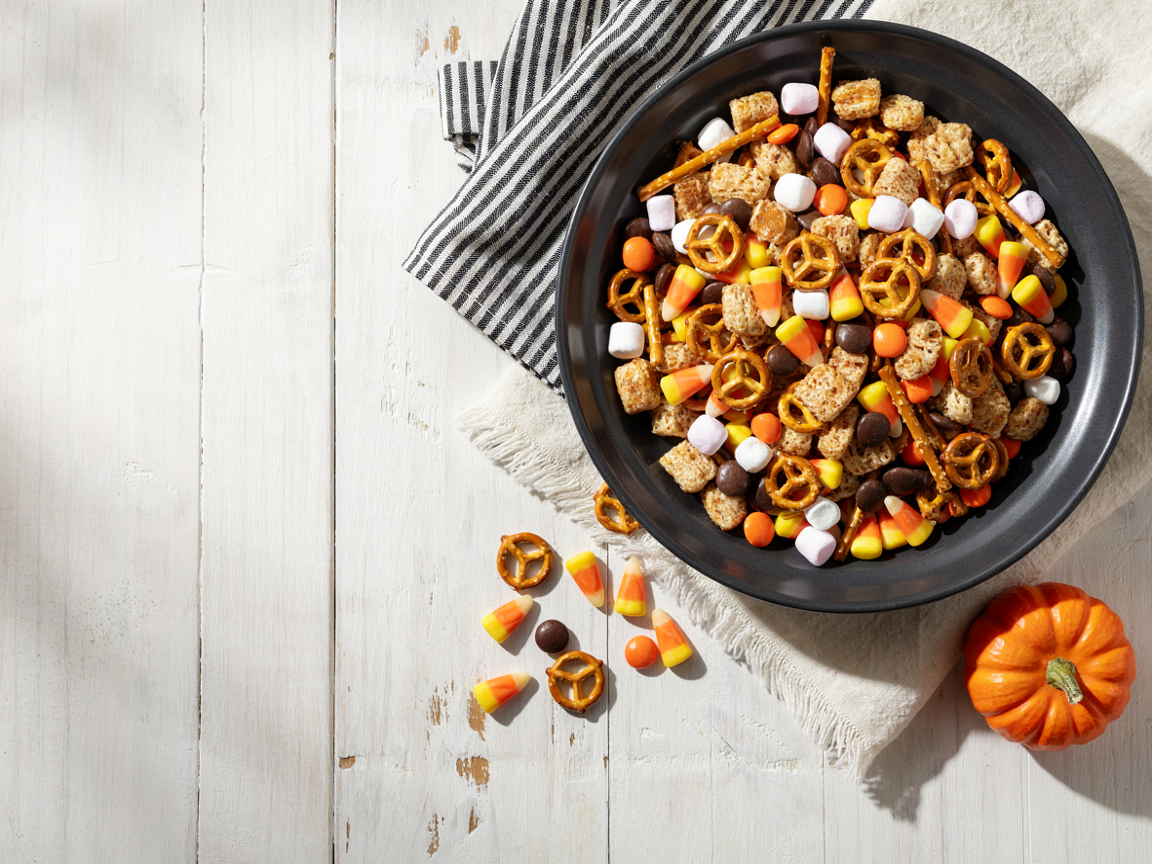 Sweet and salty Halloween snack mix in a black bowl with caramel cereal squares, pretzels, mini marshmallows, candy corn and orange and black candies