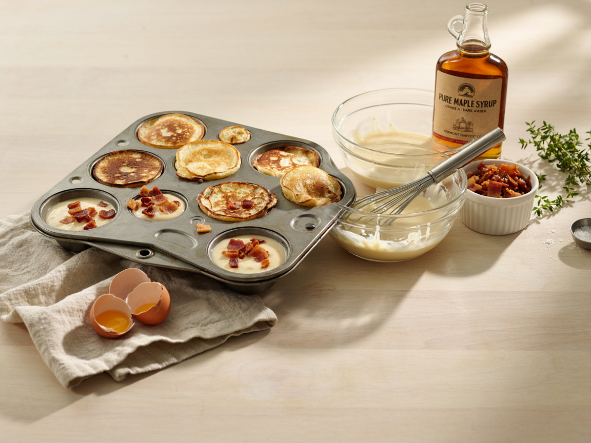 A 12-cup muffin tin with half the cups filled with pancake batter topped with bacon and half baked golden, next to a bowl of batter, a bowl of crumbled bacon and maple syrup