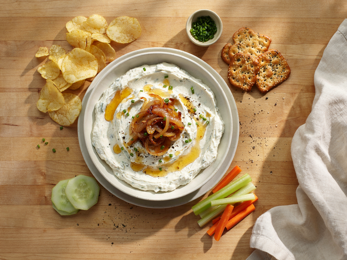 Overhead view of a ceramic bowl of caramelized onion cottage cheese dip: thick, creamy white whipped dip streaked with jammy golden-brown caramelized onions, a spoonful of onions and snipped chives on top, surrounded by kettle-cooked potato chips, celery and carrot sticks, cucumber rounds, pretzel crisps and crackers on a wooden board with a linen napkin