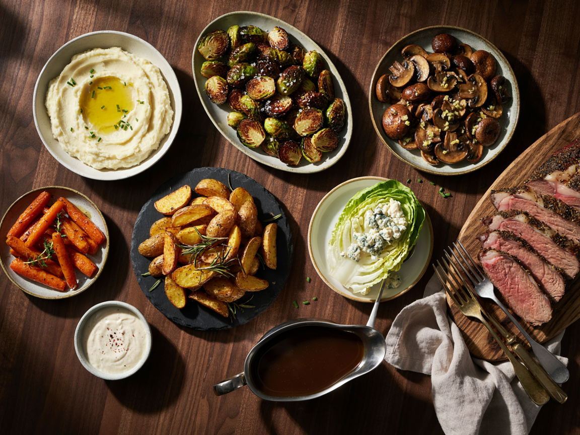 Prime rib side dishes from above on a wooden table: a bowl of creamy mashed potatoes with a pool of melted butter and chives, a platter of charred roasted Brussels sprouts, a dish of garlic butter roasted mushrooms, honey glazed carrots with thyme, crispy golden roasted potatoes with rosemary, a wedge salad with blue cheese dressing and beef bacon, a small bowl of horseradish sauce and a gravy boat of jus, with slices of rosy prime rib on a board at the edge