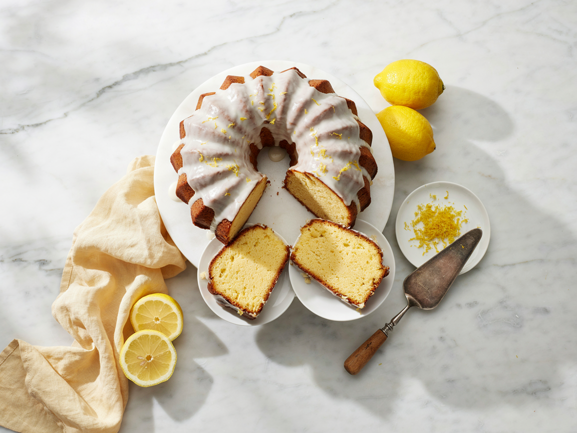 Sugar free lemon bundt cake on a white cake stand with a white lemon glaze dripping down the ridges, two slices cut and laid in front showing a soft pale yellow crumb, fresh lemons and lemon zest beside it