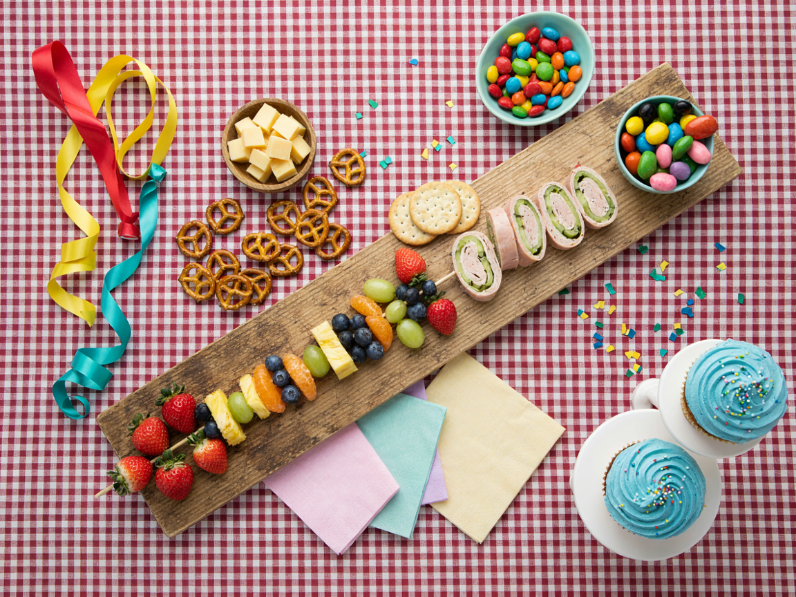 A kids birthday grazing table from above on red and white gingham: rainbow fruit skewers of strawberry, mandarin, pineapple, green grape, blueberries and red grape laid diagonally on a long wooden board, light blue bowls of candy-coated chocolates, cheddar cubes, mini pretzel twists, round crackers, rolled turkey bites, a stand of cupcakes with sky-blue frosting, red, yellow and teal balloons at the edge, bright daylight