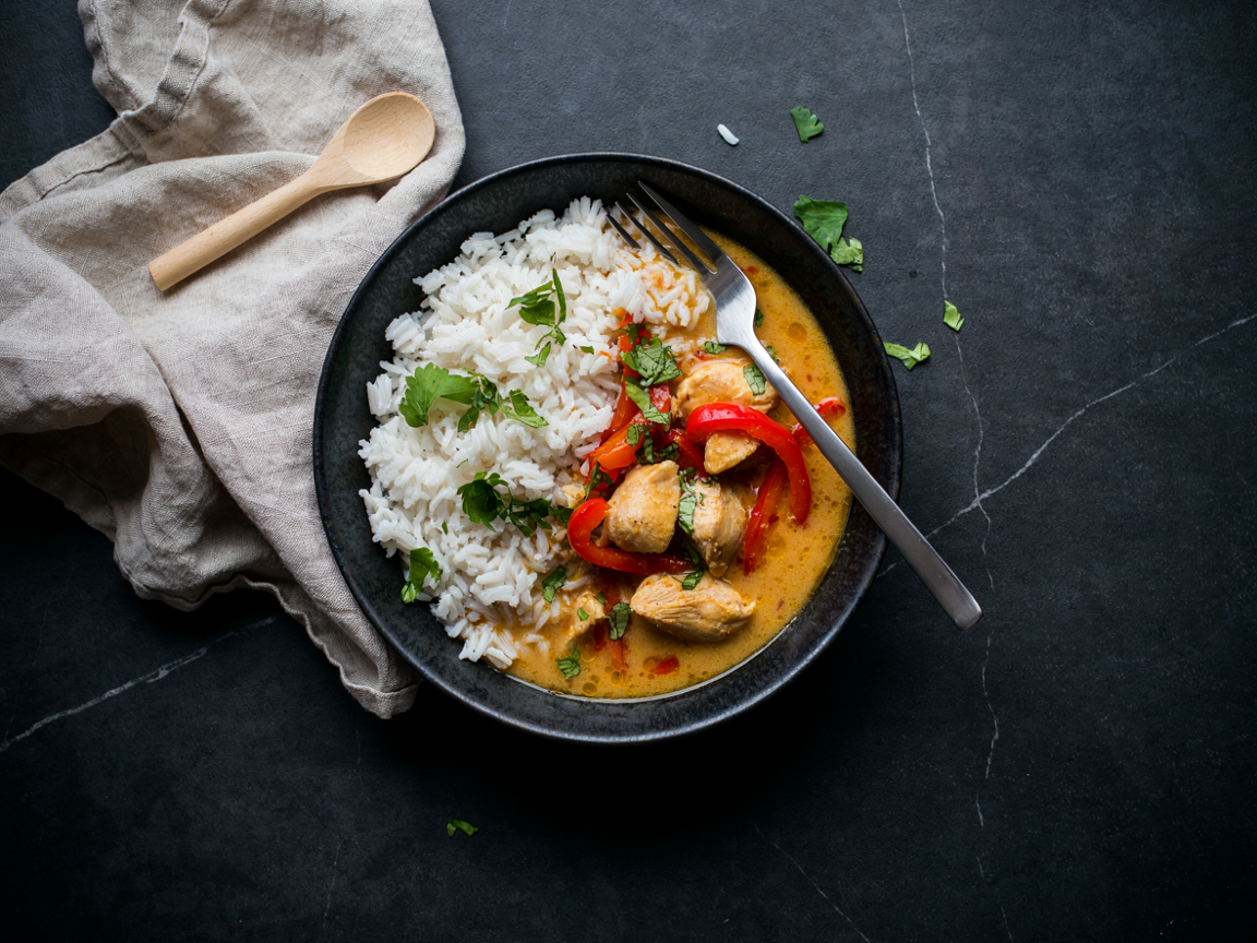 A dark charcoal ceramic bowl filled half with fluffy white basmati rice and half with creamy golden-orange coconut curry, tender chunks of chicken and glossy red bell pepper strips in a thick sauce, chopped cilantro scattered on top, a fork resting in the bowl, on a dark slate surface with a linen napkin, soft natural light, overhead food photography