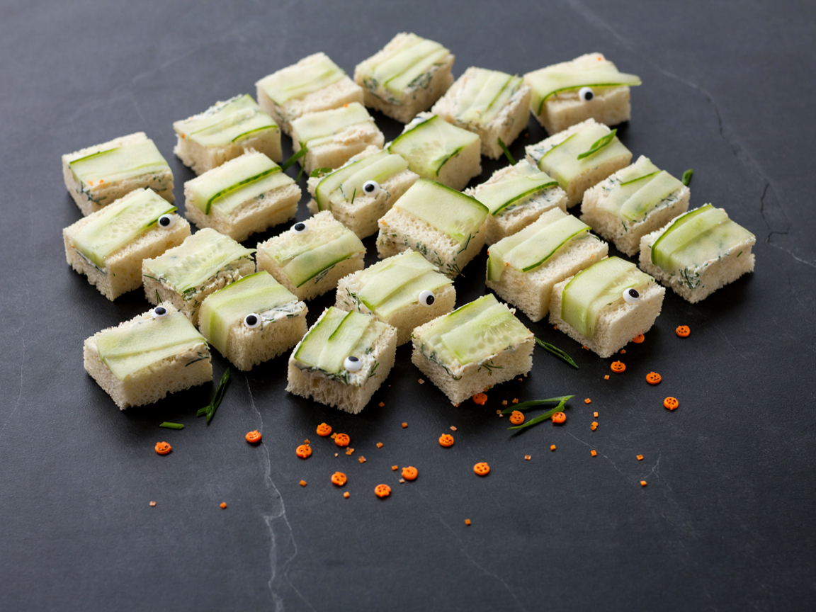 Rows of small crustless white-bread tea sandwiches filled with herby cream cheese, each wrapped in thin pale-green cucumber ribbons like mummy bandages with two candy eyes peeking out between the ribbons, on a black slate board scattered with tiny orange jack-o-lantern sprinkles, seen from above at a slight angle