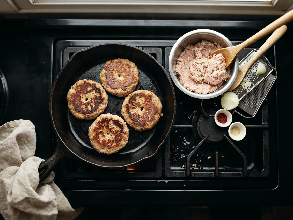 A cast-iron skillet on a stovetop from above with four turkey burger patties searing, each with a small dimple in the center, golden crusts forming at the edges, beside it a bowl of raw ground turkey mixed with grated onion and spices, a box grater with grated onion, small bowls of smoked paprika and garlic powder, warm kitchen light