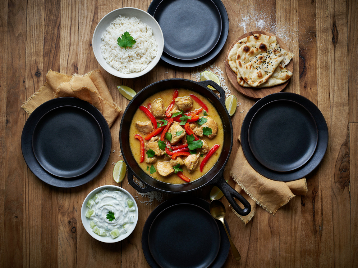 A weeknight dinner table for four from above: a large skillet of golden coconut chicken curry with red peppers and cilantro, a bowl of fluffy basmati rice, warm naan bread folded on a board, a small bowl of cucumber raita, lime wedges and four dark plates on a wooden table, warm evening light, overhead food photography
