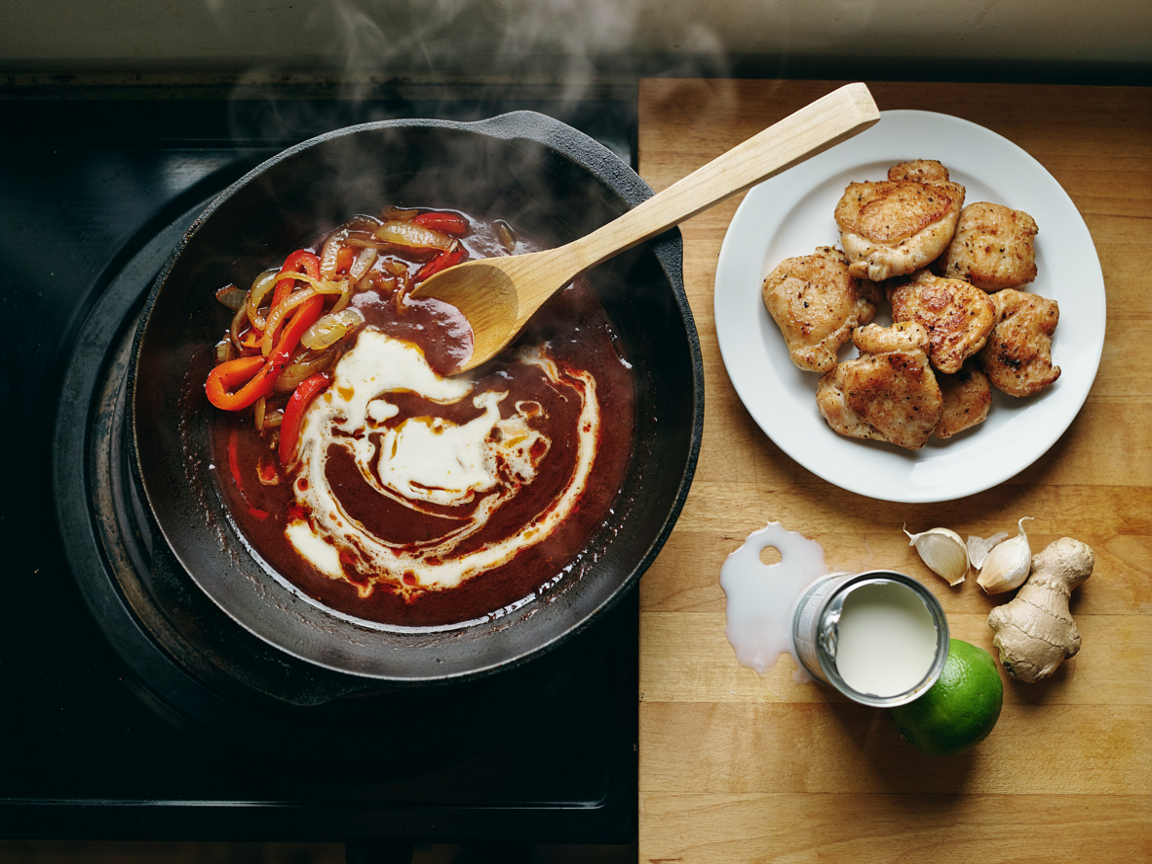 A deep skillet on a stovetop from above: dark red curry paste frying with thick white coconut cream, tiny beads of red oil appearing at the edges, softened onion and red pepper strips pushed to the side, a wooden spoon, a plate of golden-seared chicken pieces, an opened can of coconut milk, garlic, ginger and lime on the counter, warm kitchen light
