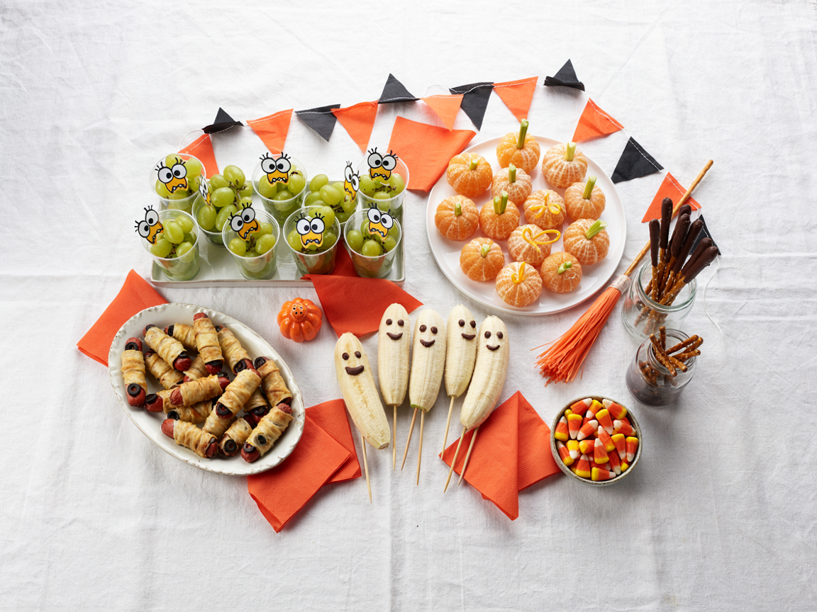 A classroom Halloween party snack table from above: a tray of clear green grape cups with cartoon monster faces, a plate of peeled tangerines with celery stems as pumpkins, mummy-wrapped mini hot dogs in golden pastry on a platter, banana halves with chocolate chip faces on sticks, chocolate-dipped pretzel rod broomsticks in a jar, orange paper napkins and black and orange paper garland on a white tablecloth, bright even light