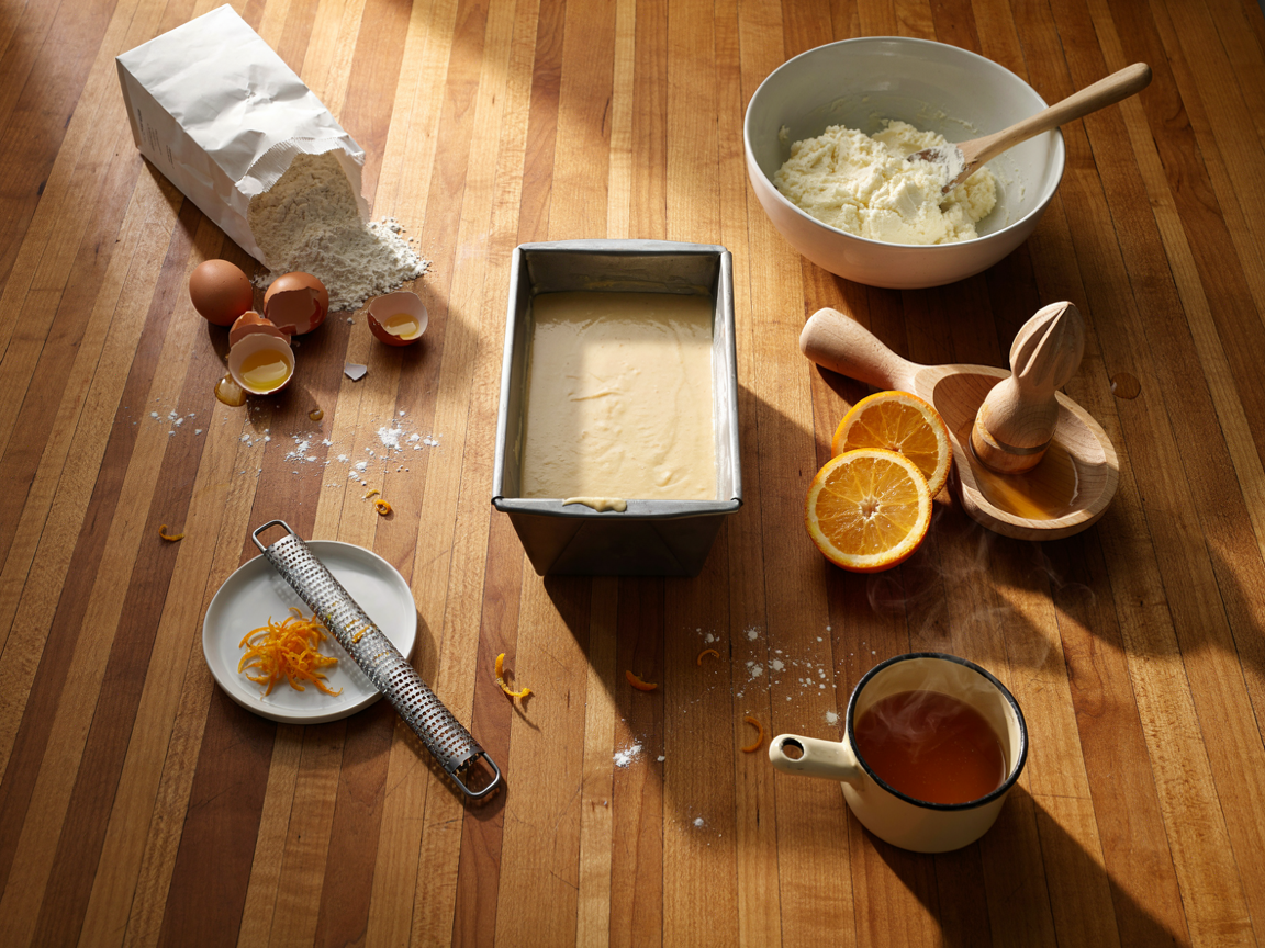 A kitchen counter from above: a loaf pan of golden pound cake batter just poured, a bowl of creamed butter and sugar, a microplane with fresh orange zest, halved oranges and a juicer, a small saucepan of orange syrup, eggs and a bag of flour, warm kitchen light