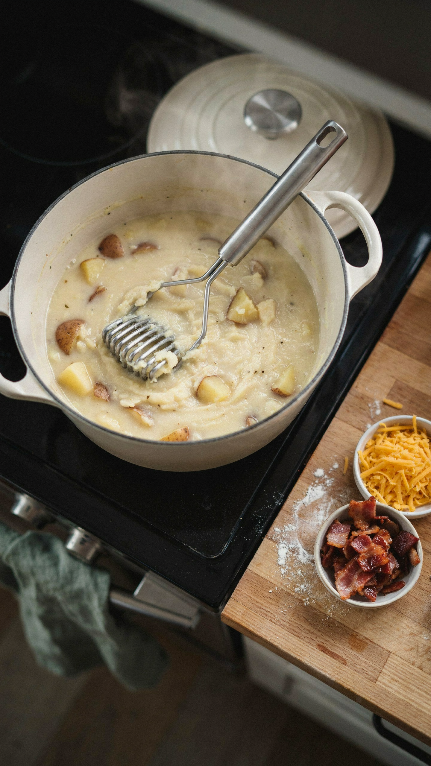 A large enameled Dutch oven on the stove full of creamy potato soup with chunks of russet potato, a stainless potato masher resting in the pot where some potatoes have been crushed into the thick base, steam rising, a bowl of shredded cheddar and a dish of crumbled beef bacon on the counter beside it, overhead angle, warm kitchen light