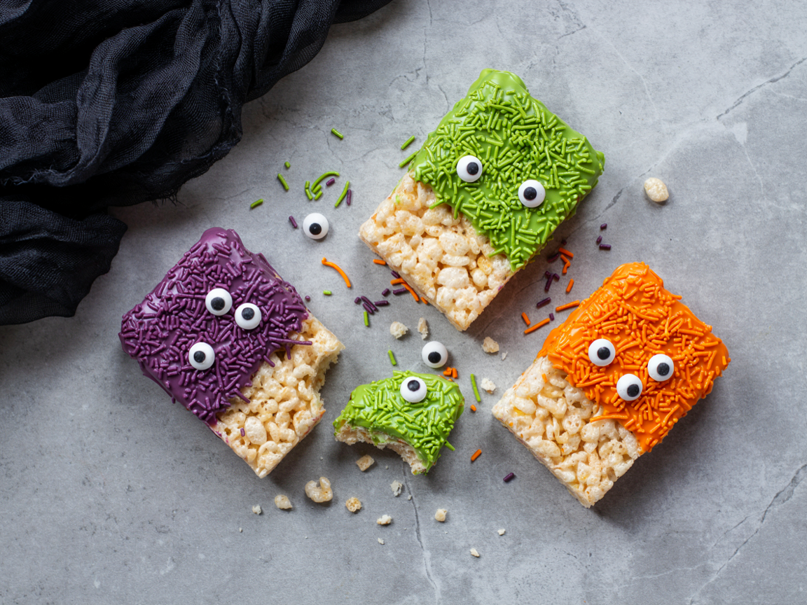 Rectangles of crispy rice cereal treats seen from above, the top third of each dipped in lime-green, grape-purple or pumpkin-orange candy coating and covered in matching long sprinkles like shaggy monster hair, with two or three candy eyes of different sizes on each, scattered on a grey stone surface with black tulle in one corner and colored sprinkles around