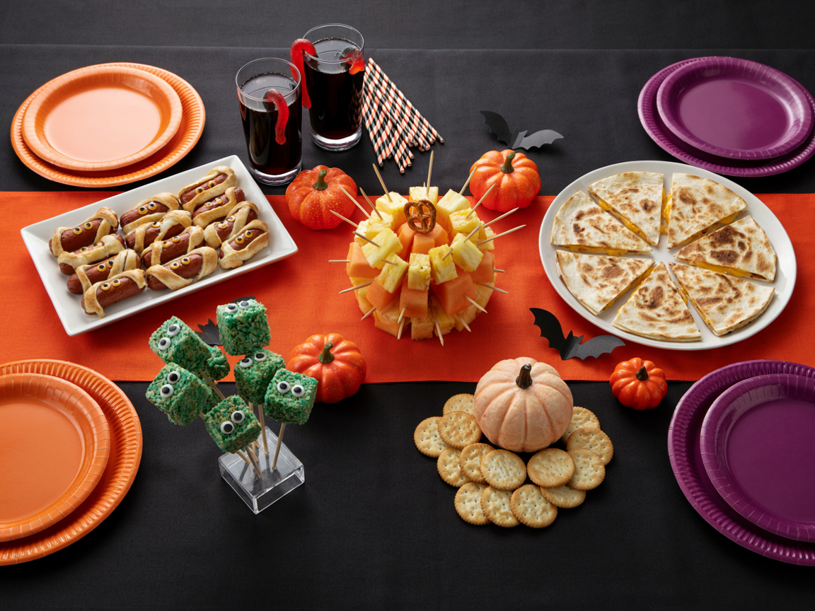 Kids Halloween party food table with the fruit skewer pumpkin in the center, mummy hot dogs, jack-o-lantern quesadillas, green monster cereal pops, a pumpkin cheese ball and glasses of black lemonade