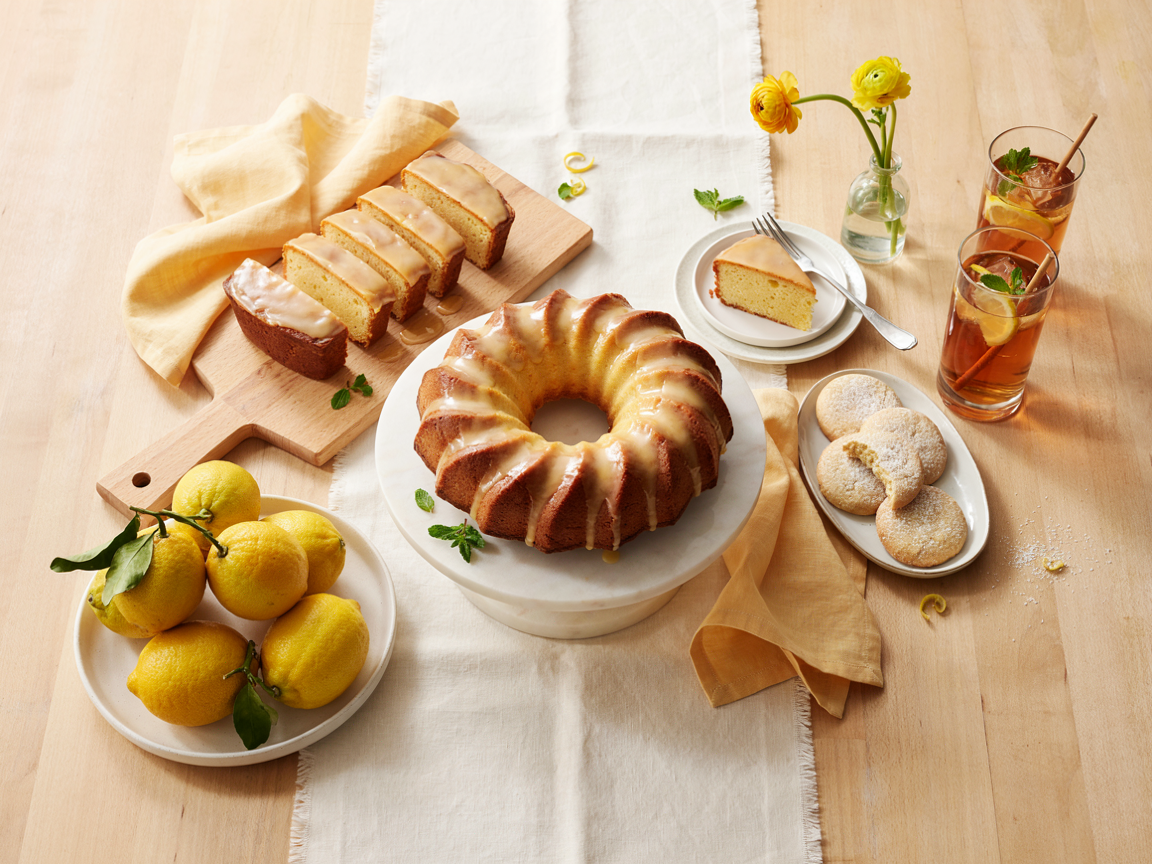 Overhead dessert table with a glazed sugar free lemon bundt cake in the center, a slice on a plate with a fork, a plate of lemon cookies, a loaf cake, glasses of iced tea with lemon slices, fresh lemons and a pale yellow linen