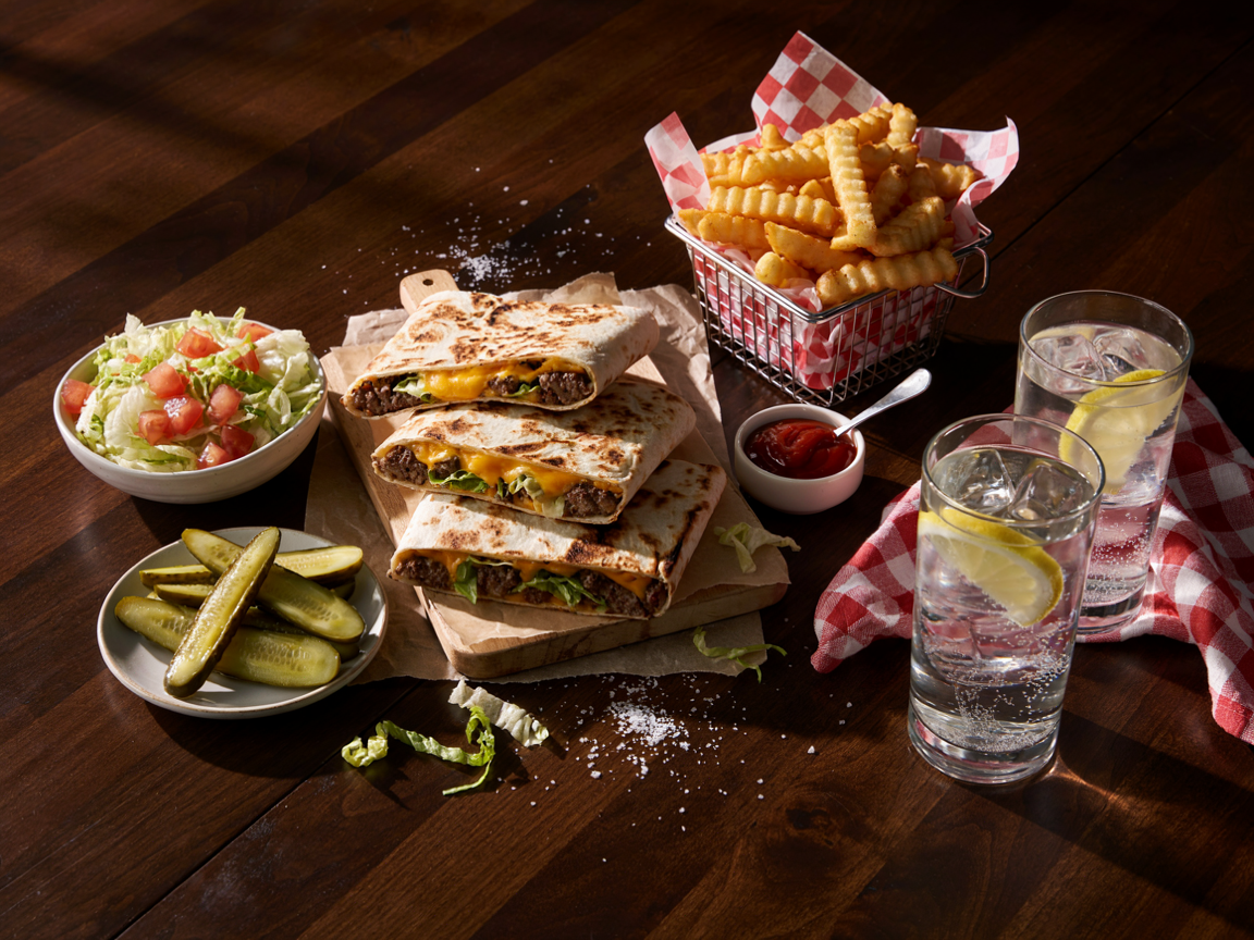 Burger night table from above: four cheeseburger burritos cut diagonally and stacked on a parchment-lined board, a basket of golden fries, a bowl of pink burger sauce, dill pickle spears, a small bowl of shredded lettuce and diced tomato, and two glasses of iced lemonade on a wooden table