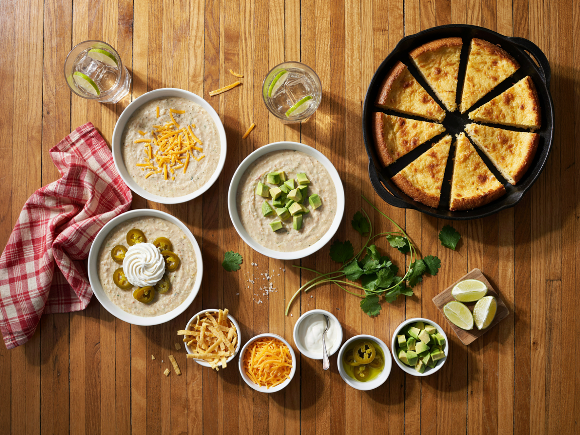 Table spread of white chicken chili bowls with a skillet of cornbread and small bowls of toppings: tortilla strips, avocado, cheese, jalapenos and lime wedges