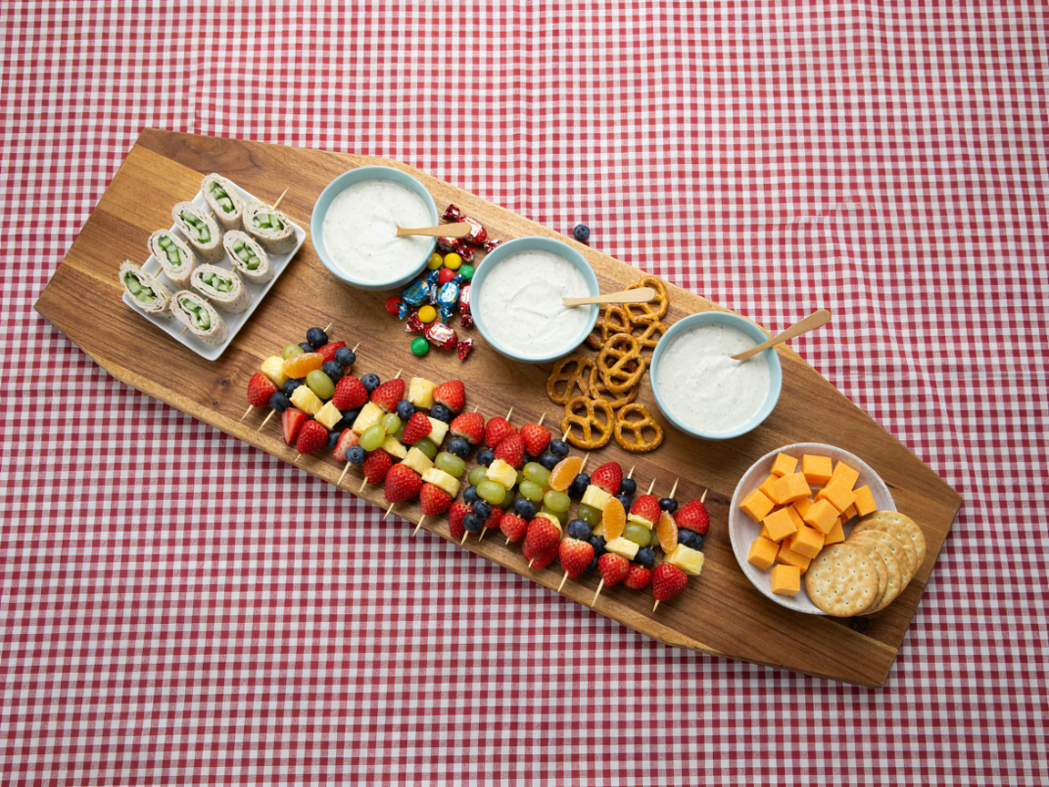 A kids grazing table being set up, seen from above: a long wooden board on red gingham with light blue bowls of ranch, candy and pretzels already placed, a row of rainbow fruit skewers along the front, a tray of rolled turkey bites and a bowl of cheddar cubes waiting beside the board, crackers still stacked in their sleeves, bright daylight