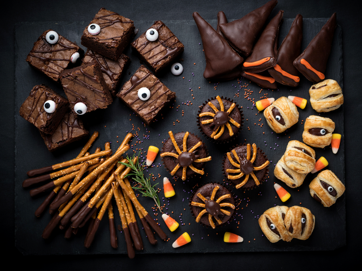 A Halloween party snack board on black slate from above: fudgy brownie squares with a chocolate drizzle and mismatched candy eyeballs, mummy-wrapped pastry bites with golden strips and candy eyes, chocolate-dipped pretzel rod broomsticks, chocolate witch hat cookies with orange frosting bands, chocolate cupcakes with pretzel-leg spiders, scattered candy corn and orange and purple sprinkles, dark moody light with warm highlights