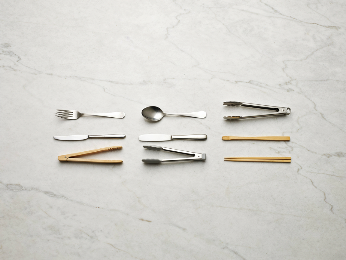 Overhead flat lay on marble showing a metal fork, knife, spoon and metal tongs in the top rows and bamboo tongs, silicone tongs and wooden chopsticks below