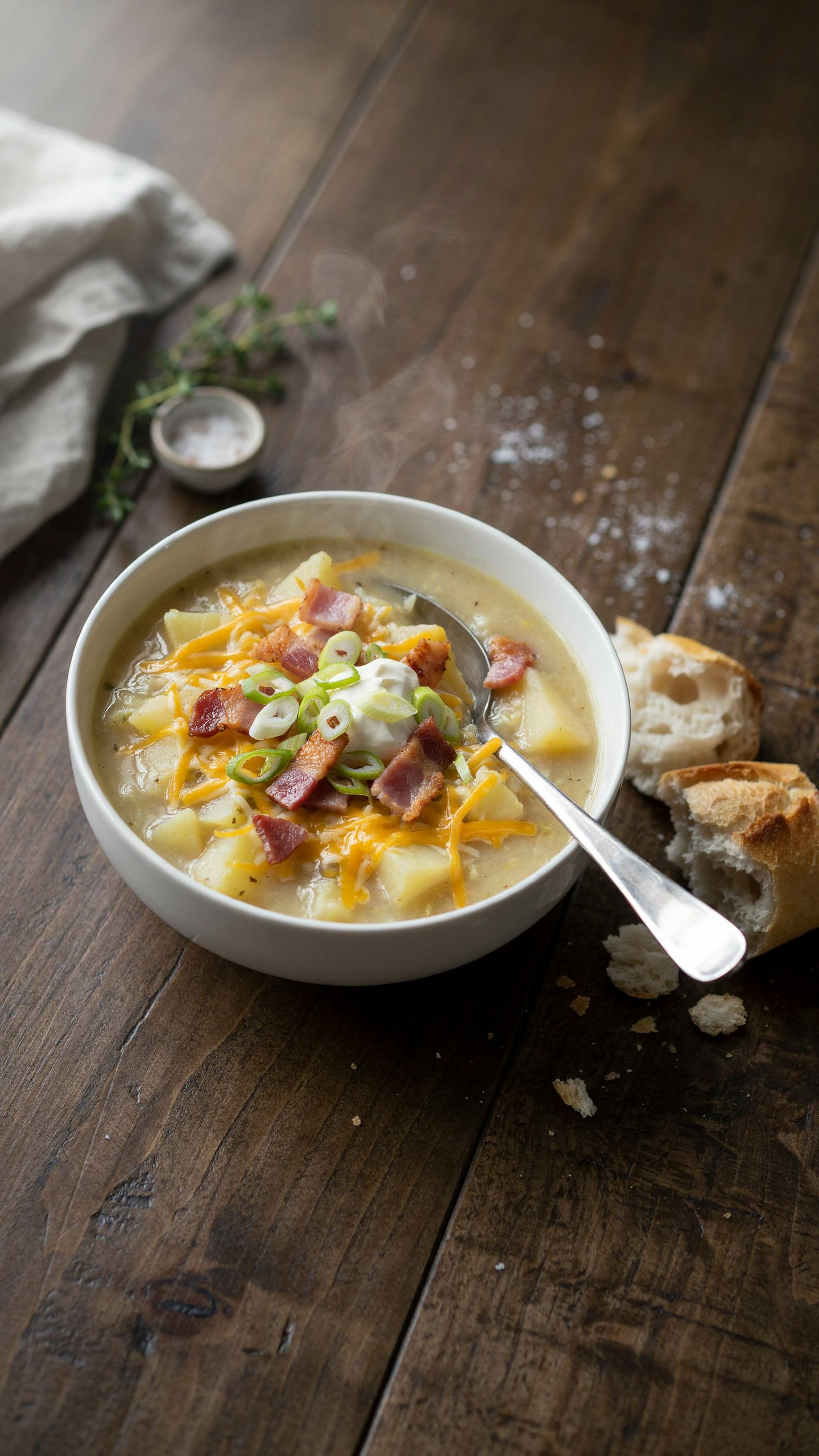 A deep white ceramic bowl of thick creamy loaded baked potato soup, pale gold with visible chunks of potato, topped with melted shredded cheddar, crumbled crispy beef bacon, a dollop of sour cream and sliced green onions, a spoon resting in the bowl, a torn piece of crusty bread beside it on a rustic wooden table, warm natural light
